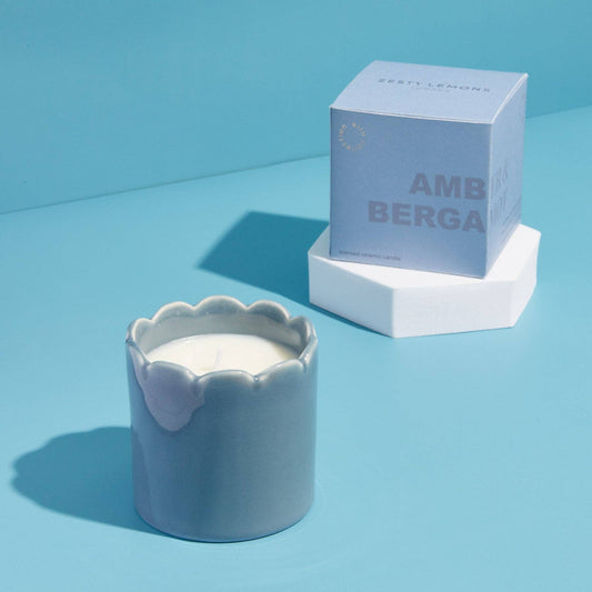 Candle in a scalloped container with a box labelled 'Amber and Bergamot' on a blue background