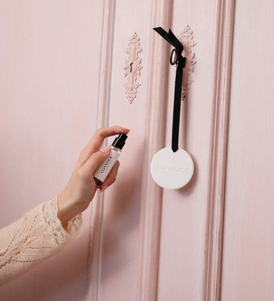 Hand holding a room mist bottle spraying a white circular disc hanging on a black ribbon on a pink wardrobe.