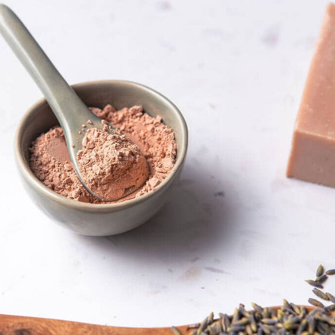Small bowl with pink powder, a spoon, a bar of soap, and lavender flowers on a light surface.