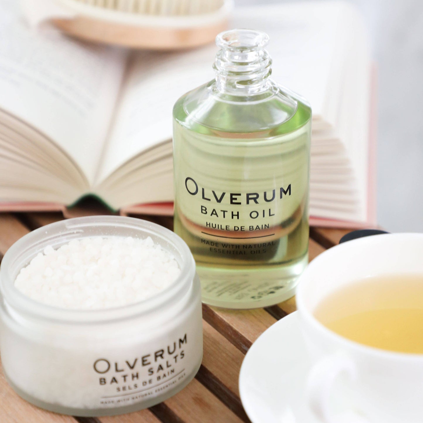 Olverum bath oil and bath salts on a wooden surface with a book and cup in the background.
