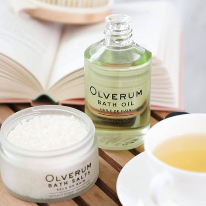 Olverum bath oil and bath salts on a wooden surface with a book and cup in the background.