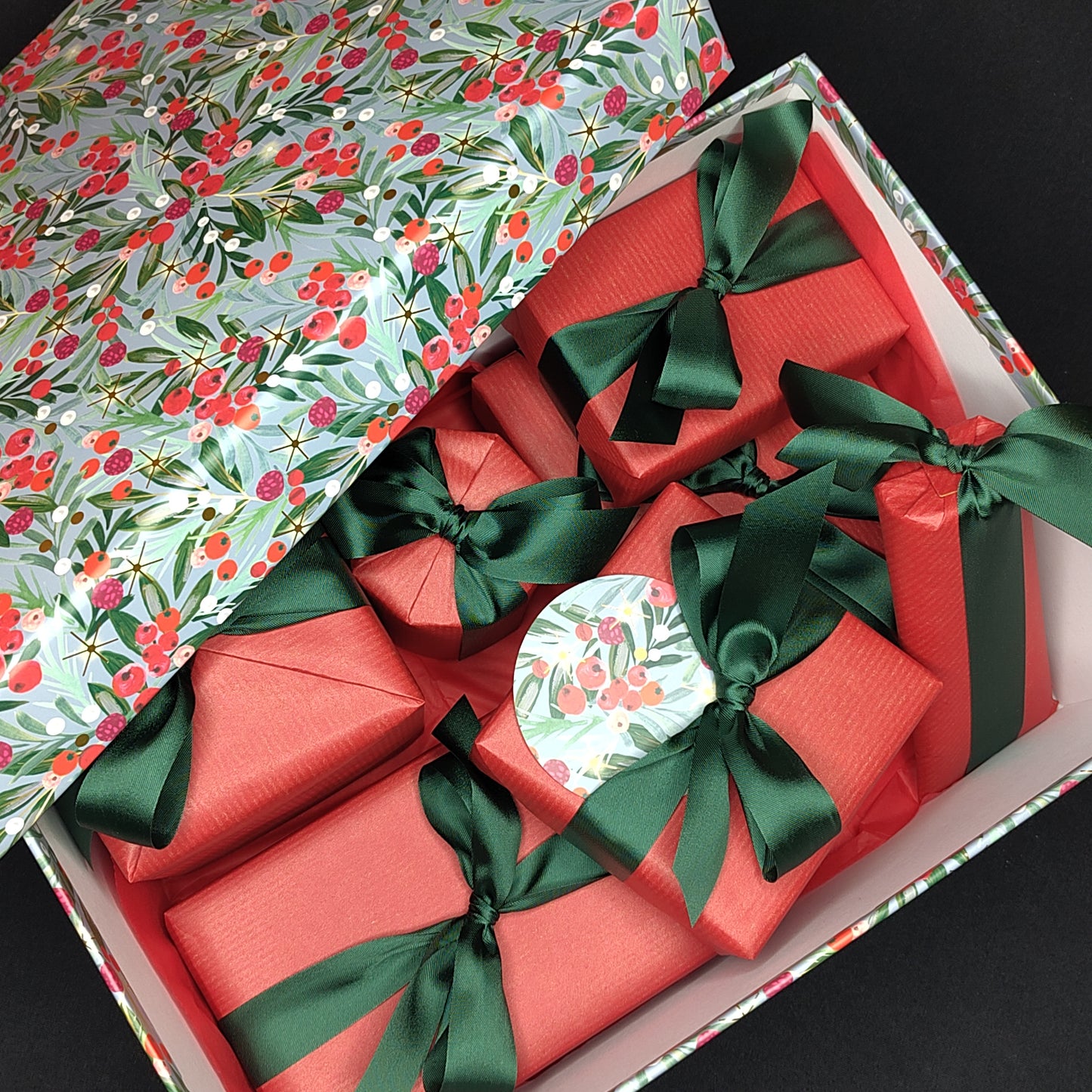 A gift box patterned with blue, green leaves and red berries filled with presents wrapped in red paper with dark green ribbon.